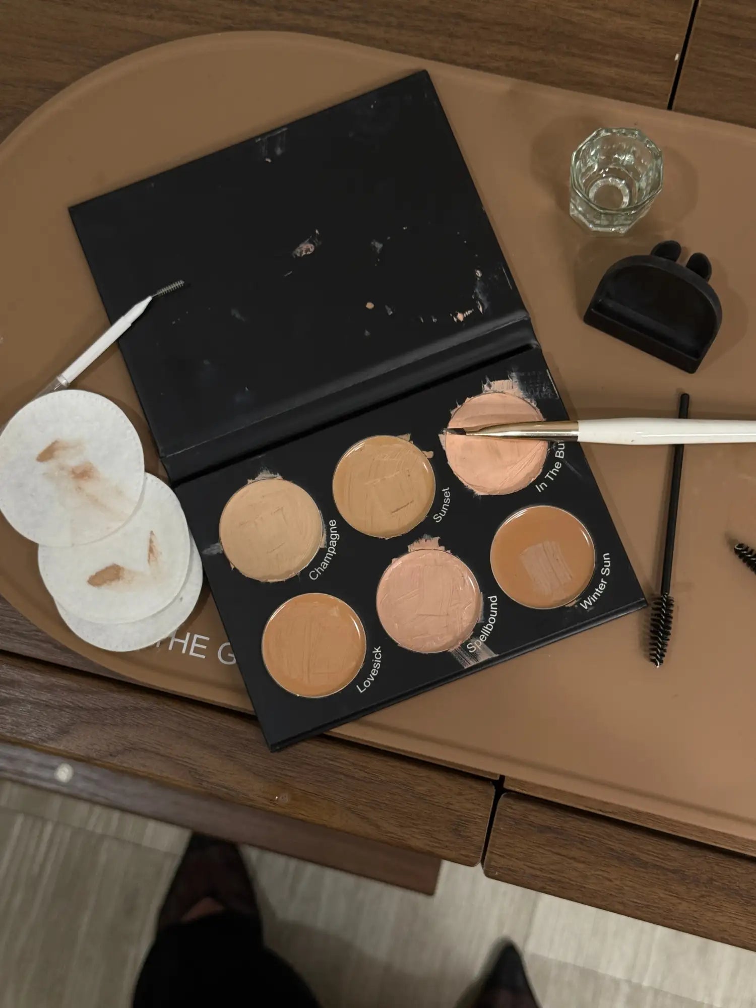 Black concealer palette with six circular pans of varying beige and peach tones, labeled ’crimson,’ ’sunset,’ ’in the sun,’ ’loveback,’ ’skylightbound,’ and ’winter sun’