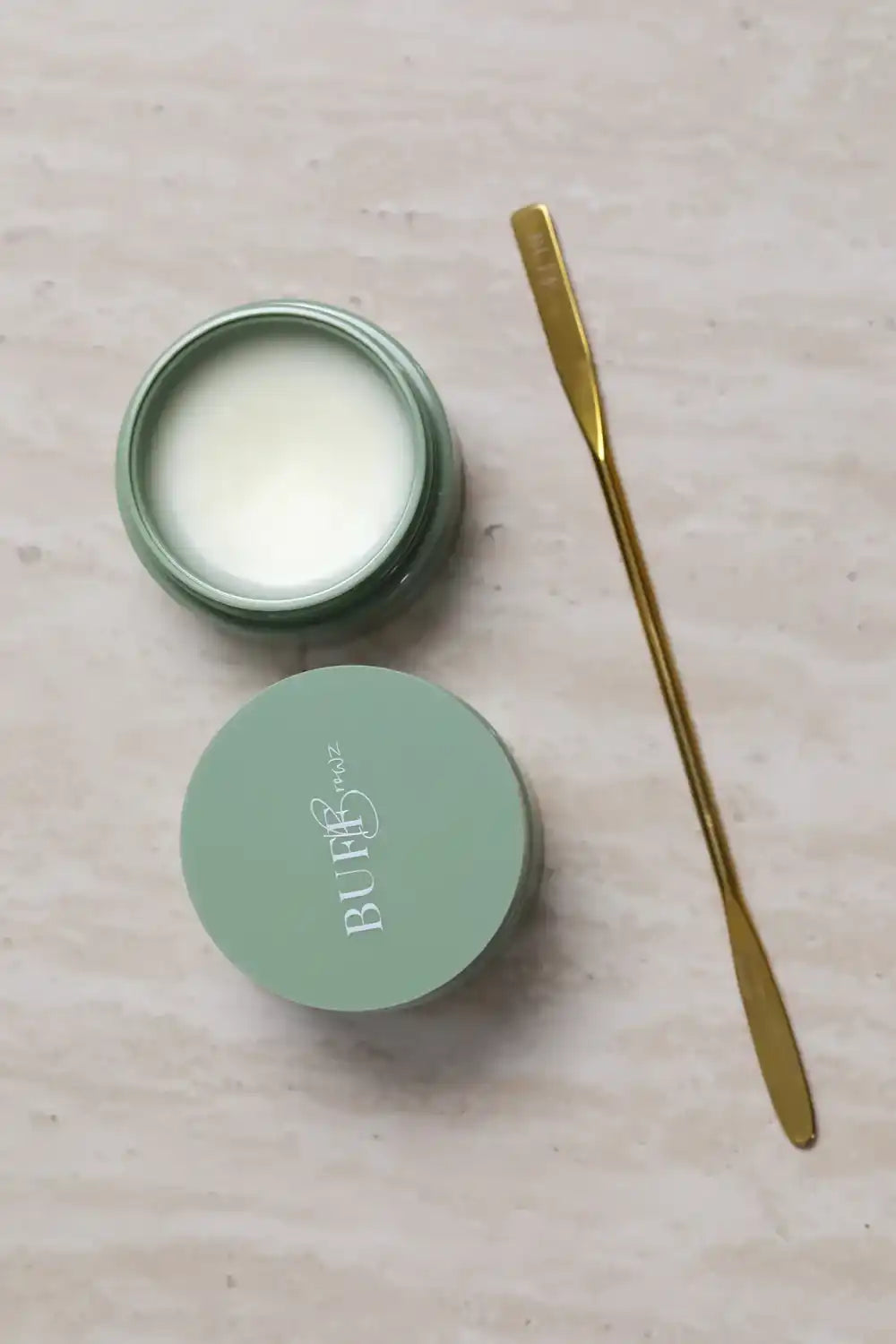 An open jar of light-colored balm with a matching lid and a gold-colored spatula.